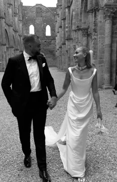 Couple portrait of bride and groom holding hands, bride in veil with bouquet beside tuxedo groom in stone ruin courtyard arches