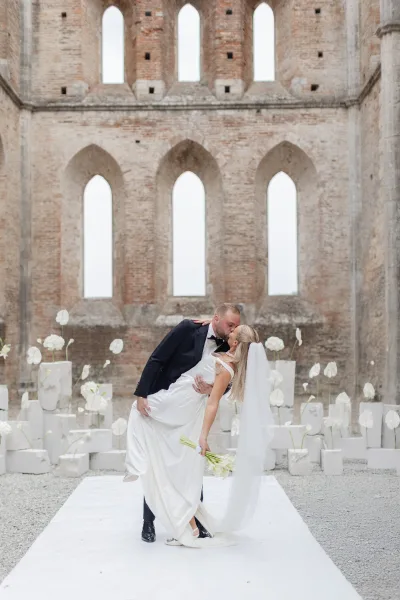 Wedding kiss as groom dips the bride in off-the-shoulder dress and long veil, holding a simple bouquet in brick church ruins aisle