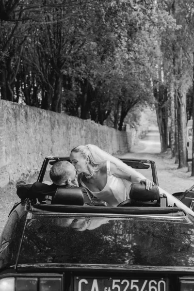Wedding kiss portrait of newlyweds in convertible, bride in veil and pearl necklace leaning in as groom in suit, tree-lined road behind