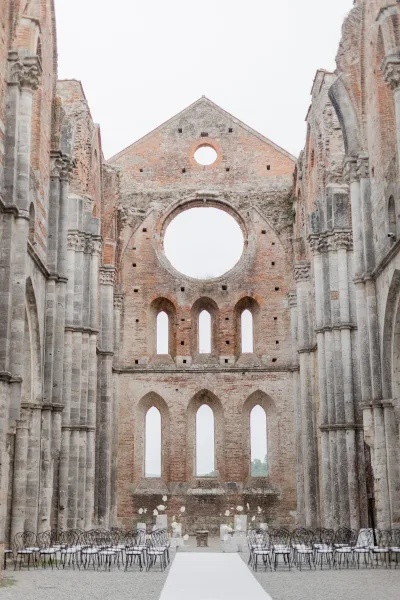 Ceremony setup with outdoor ceremony aisle and white aisle runner, wrought iron chairs and florals framed by cathedral ruins and open sky