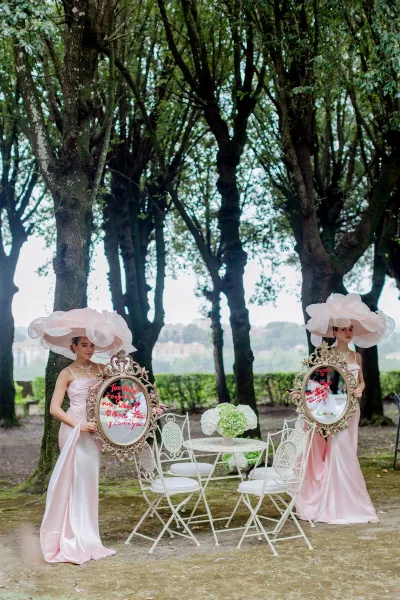Bridesmaid portrait with bridesmaids holding mirrors in blush pink satin dresses and oversized hats by a white bistro table in a tree grove
