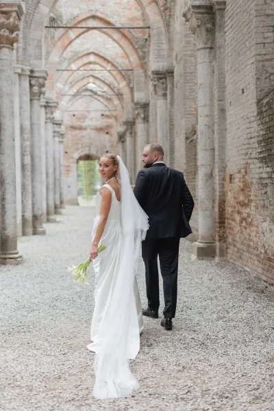 Couple portrait of bride and groom walking away, bride looking back with long veil and calla lily bouquet under stone archway