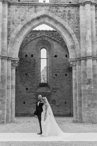 Couple portrait in a black and white wedding portrait under a stone archway, bride in veil and sheath dress with groom in tuxedo by brick wall