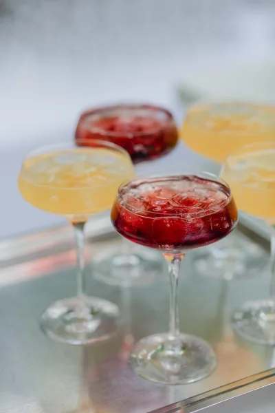Wedding cocktails in coupe glasses with ice cubes, featuring berry red and citrus yellow drinks on a metal tray against a white backdrop