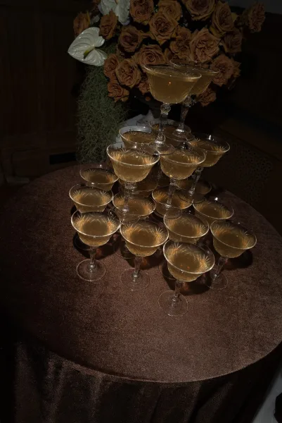 Champagne tower setup with stacked coupe glass champagne tower on a round table, paired with rose florals and greenery in a moody interior setting
