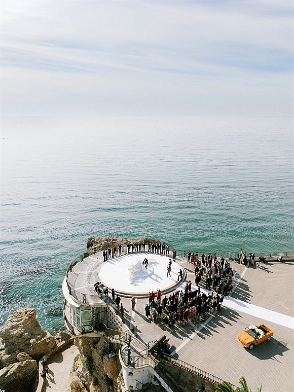 Outdoor wedding ceremony on a circular platform with a white aisle runner and chairs overlooking an ocean and rocky cliffside terrace