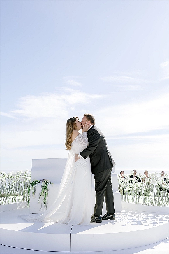 Wedding kiss portrait of bride and groom kiss on a modern white ceremony platform, off-shoulder gown and train, guests and sky behind