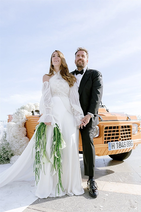 Couple portrait of bride and groom holding hands beside a vintage car, bride in sleeves gown with cascading greenery bouquet under blue sky