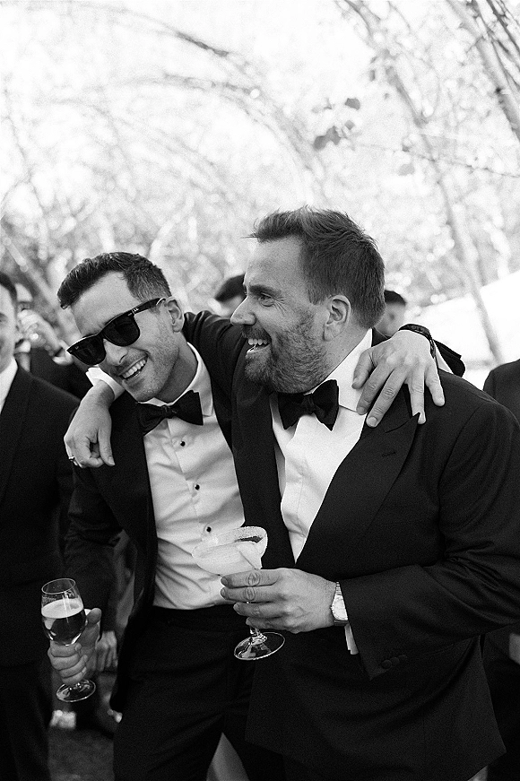 Groomsmen portrait with tuxedos and bow ties, laughing in sunglasses while holding cocktail and wine glasses among trees and guests