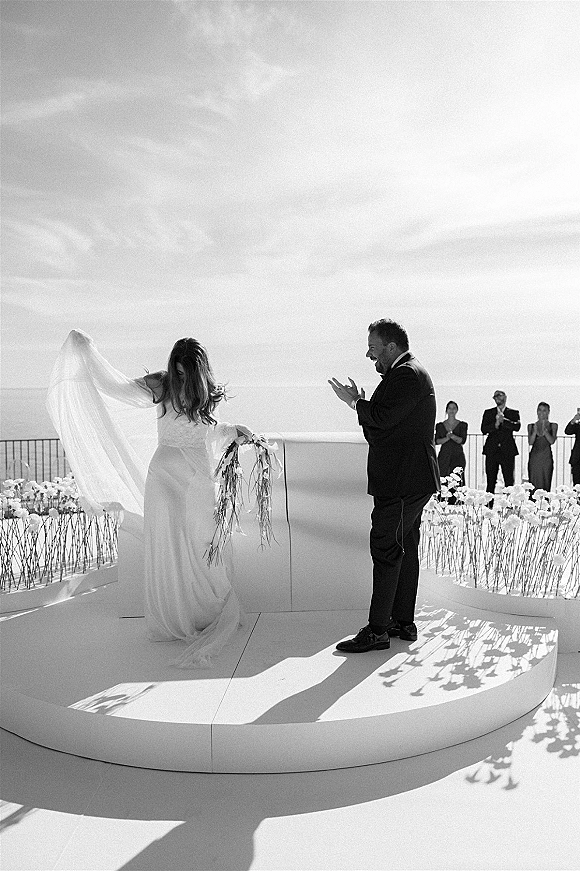 Ceremony moment as bride walks down aisle at outdoor wedding ceremony, veil flowing, bouquet ribbons trailing, guests by oceanfront platform