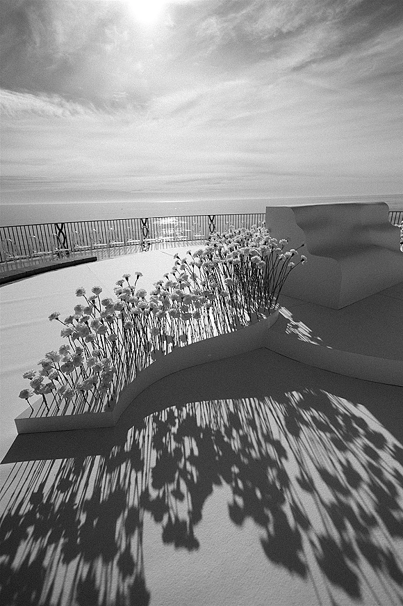 Ceremony setup with oceanfront ceremony setup features white florals lining a curved platform aisle, clear chairs and bench seating on a terrace overlooking the horizon