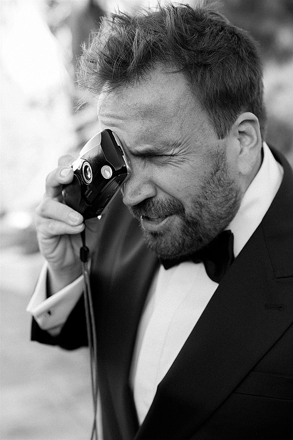Groom portrait in a black tuxedo holding a compact camera by the strap, smiling on an outdoor walkway with blurred guests behind him