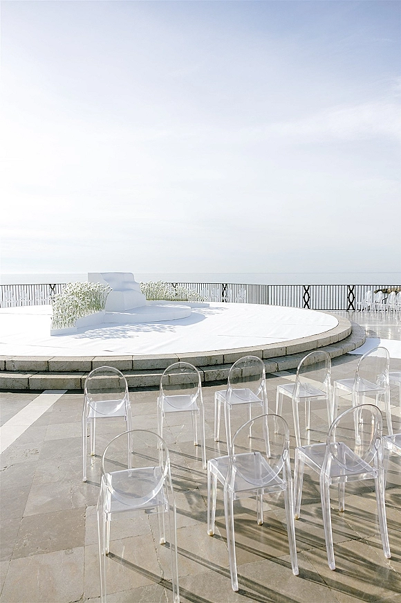 Ceremony setup for an outdoor wedding ceremony with clear acrylic chairs facing a round white stage, white florals, and ocean view backdrop