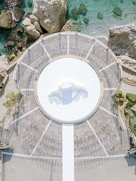 Ceremony setup with a circular wedding ceremony layout, clear chairs around a white aisle runner on a stone terrace by the oceanfront floral arch