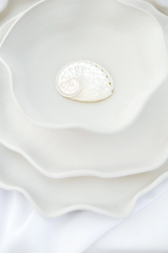 Wedding table setting with white ceramic and scalloped plates, a seashell place card, and neatly folded white napkin on a white tabletop