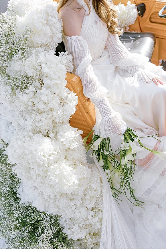 Bridal portrait of a bride in vintage car holding a white calla lily and hydrangea bouquet, seated on a leather seat by florals