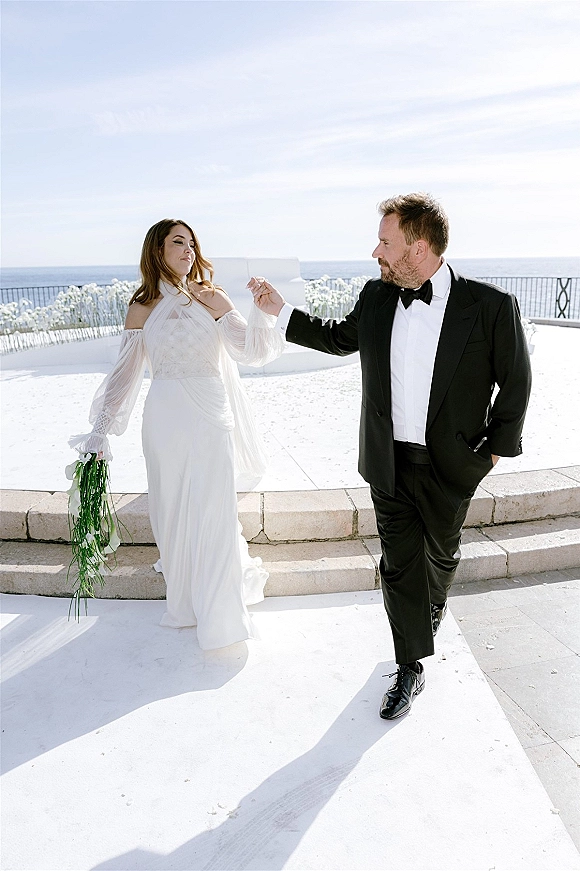 Couple portrait of bride and groom holding hands, her off-shoulder dress and calla lily bouquet on an oceanfront terrace with white florals