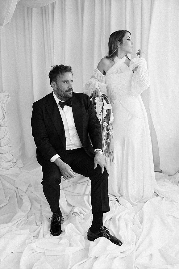 Wedding portrait of bride standing in an off-the-shoulder gown with bouquet beside seated groom in tuxedo on chair, studio draping backdrop