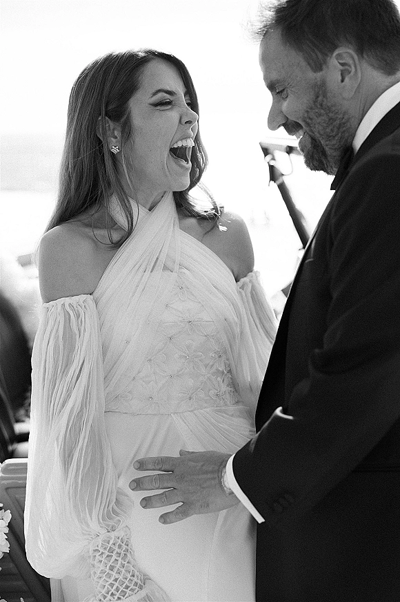Wedding couple portrait in black and white, bride laughing in an off-the-shoulder floral applique gown as groom looks on by bright windows