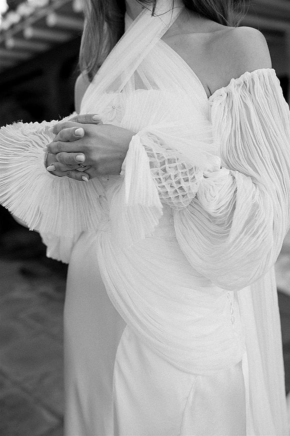 Wedding dress detail with off the shoulder wedding dress sleeves, ruched bodice, and bride’s ringed hands against blurred greenery outdoors