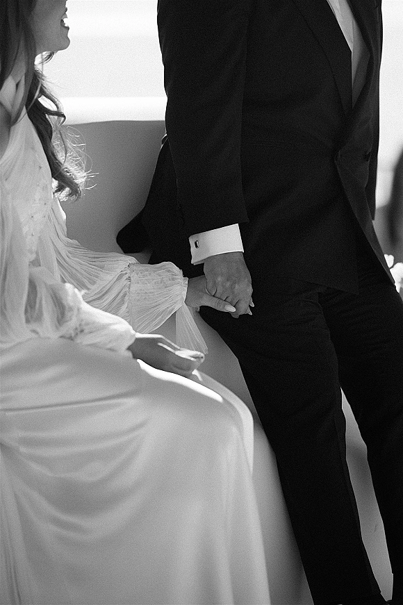 Couple holding hands in a wedding handholding photo, bride’s long-sleeve dress and groom’s tuxedo cufflinks in soft window light indoors