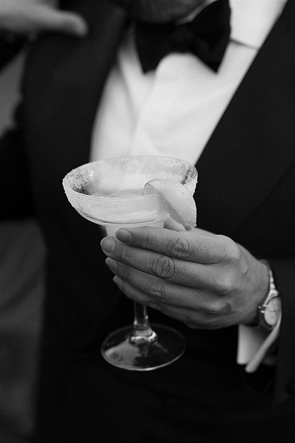 Wedding cocktail held by a groom in a tuxedo, a signature wedding drink in a salt-rim coupe glass with citrus garnish and blurred guests behind