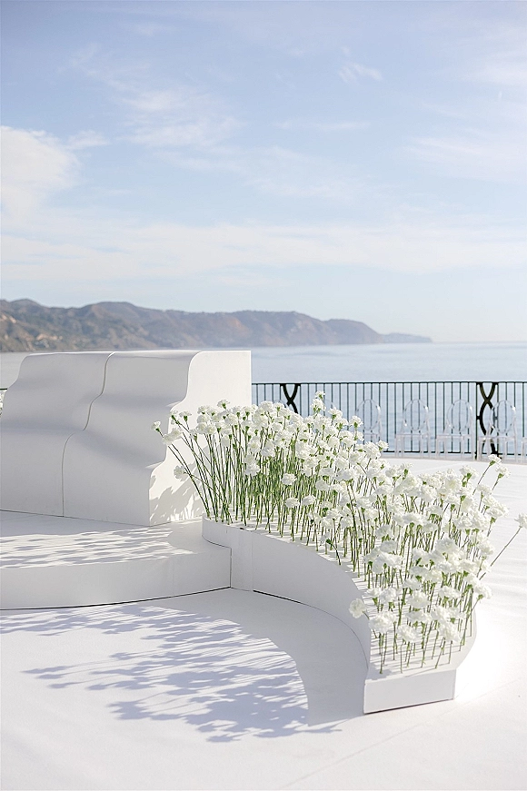 Ceremony altar with modern wedding altar styling, sculptural white backdrop and white floral meadow arrangement on a terrace with ocean views
