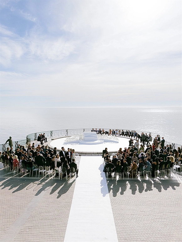 Ceremony setup with a white aisle runner leading to a circular platform altar on a stone terrace overlooking the ocean and sky