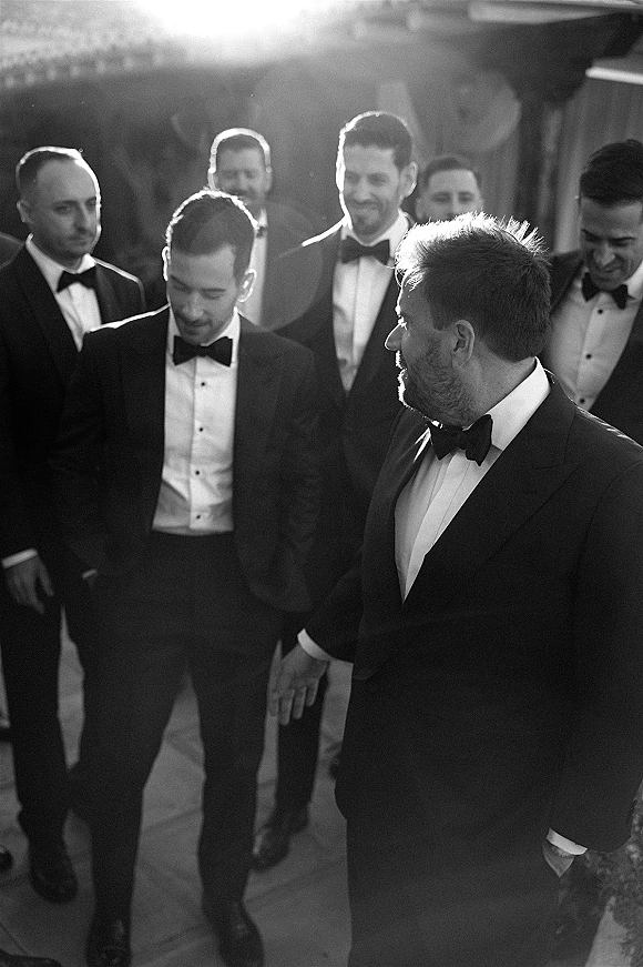 Groomsmen portrait of groom and groomsmen in tuxedos with bow ties and dress shirts, standing on an outdoor patio by a building exterior
