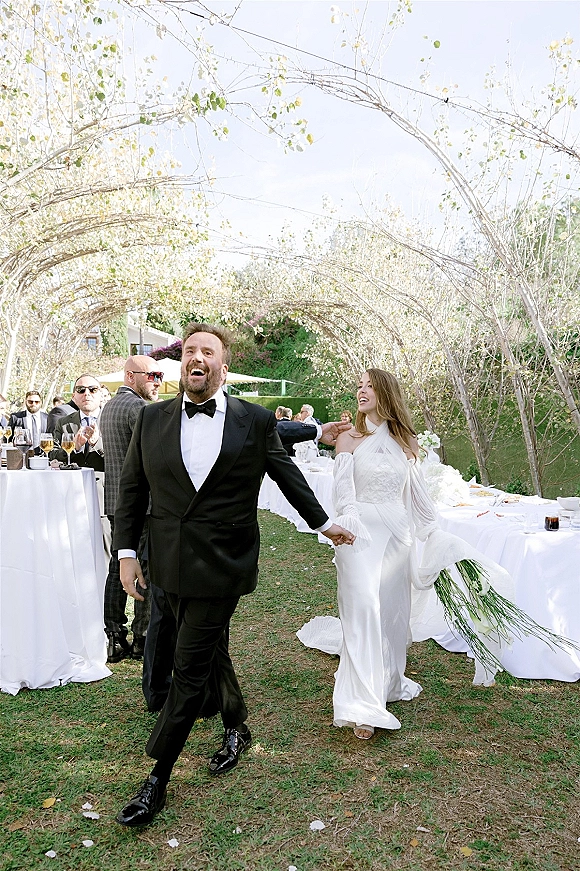 Reception entrance as bride and groom walk hand in hand past cheering guests under string lights on a garden lawn with cocktail tables