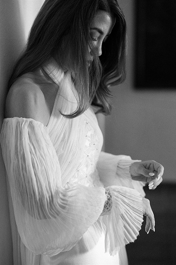 Bridal portrait in black and white of a bride looking down, adjusting her off-the-shoulder pleated wedding dress in an indoor room