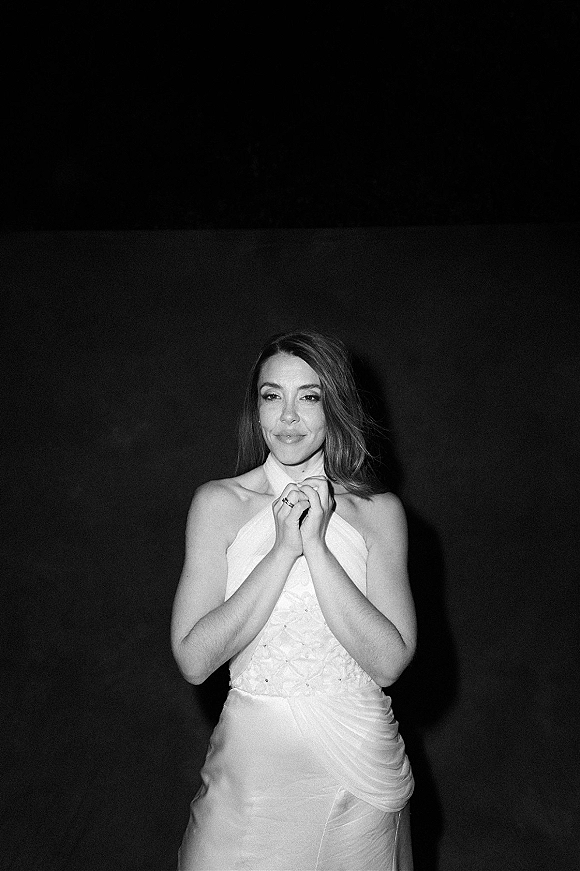 Bridal portrait of a bride in a halter wedding dress with lace bodice, hands clasped showing engagement ring against a dark wall backdrop