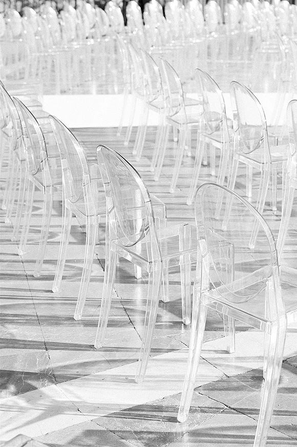 Ceremony seating with clear acrylic wedding chairs arranged in rows, sunlight casting shadows across a stone tile outdoor floor