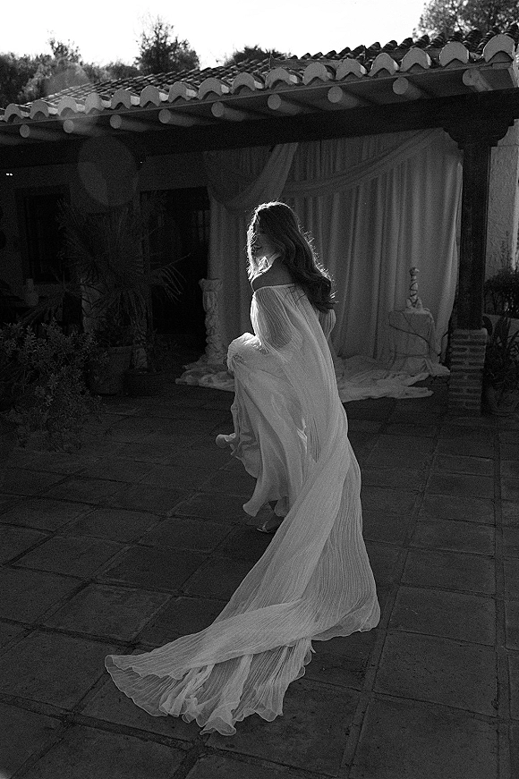 Bridal portrait of a bride walking away, looking over her shoulder as her long train flows across a stone patio by stucco walls and plants