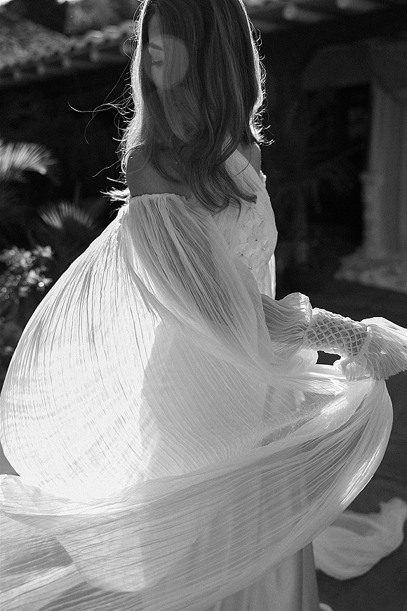 Bridal portrait in black and white of a bride twirling a pleated chiffon off-the-shoulder gown on a sunlit garden stone walkway