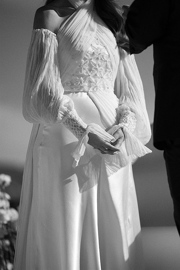 Wedding dress close-up showing off the shoulder sleeves with 3D floral applique bodice and lace cuffs against a plain wall backdrop