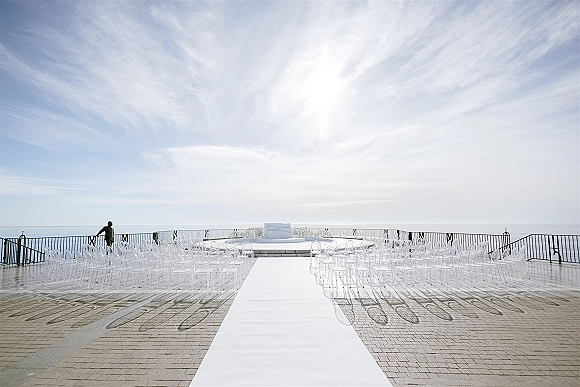Ceremony setup with a white aisle runner leading to a platform, clear chairs aligned on a stone patio with ocean horizon beyond