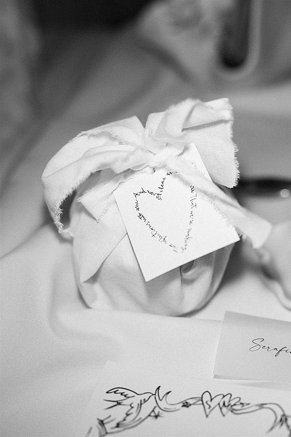 Wedding favor wrapped in fabric with a tied bow and handwritten gift tag on a tabletop beside a blurred envelope and place card