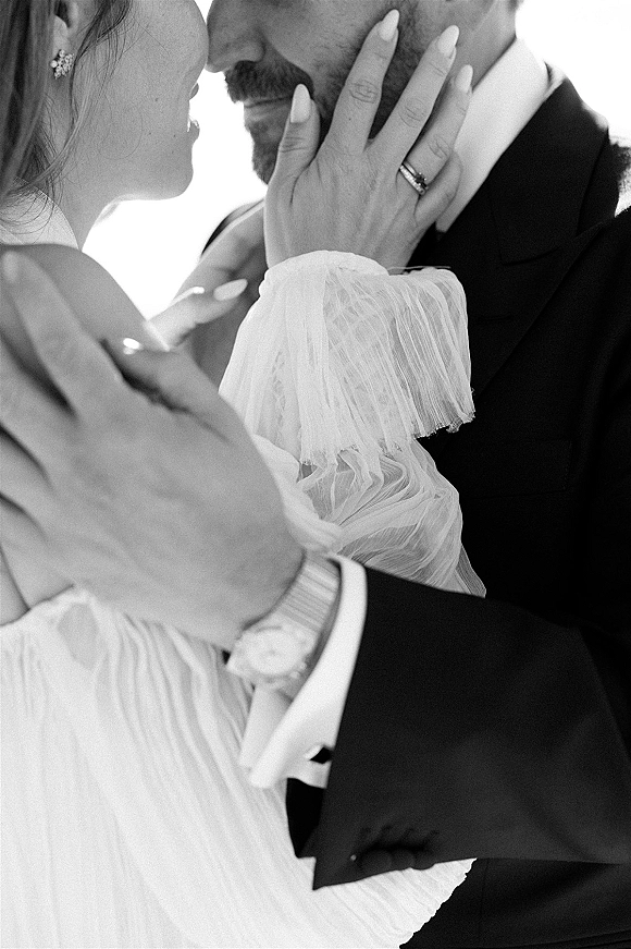 Wedding couple portrait close up as bride touches groom’s face forehead to forehead, showing wedding rings and textured long sleeve gown against bright sky