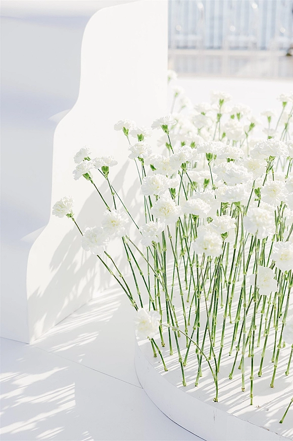 Wedding floral installation with single-stem white flowers on circular bases, creating a minimalist meadow beside white seating in window light