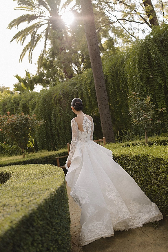 Bridal portrait of a bride walking away in a backless lace wedding dress with button details, on a sunlit garden path with hedges and palm trees