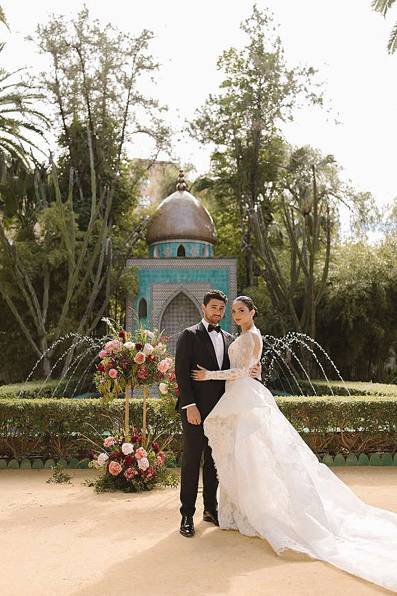 Couple portrait of bride in long sleeve lace wedding dress and groom in black tuxedo posing by a garden fountain under a tiled pavilion