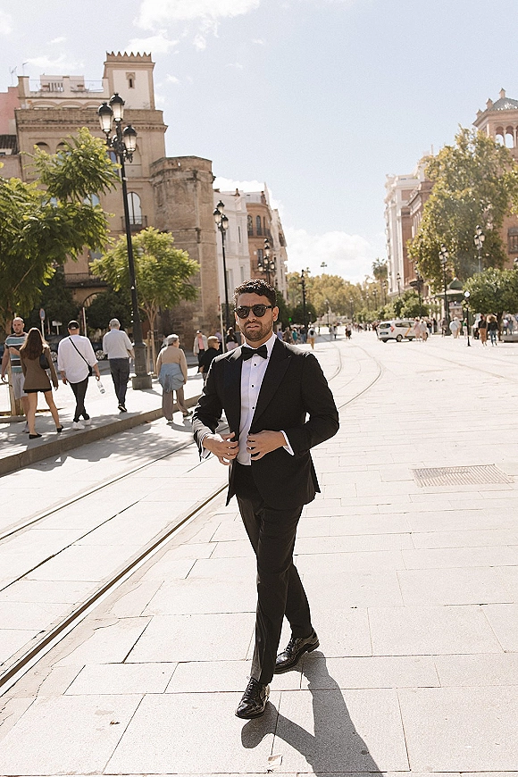 Groom portrait in a black tuxedo walking in sunglasses along a city street with historic buildings, street lamps, and tram tracks in daylight