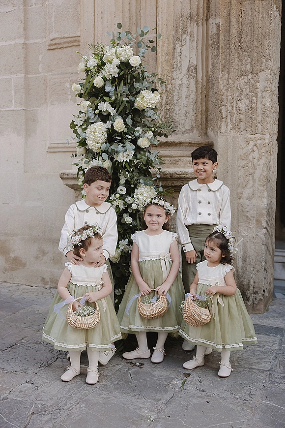 Flower girl and ring bearer in sage green dresses and suspenders hold wicker baskets with white flowers on stone steps by a facade