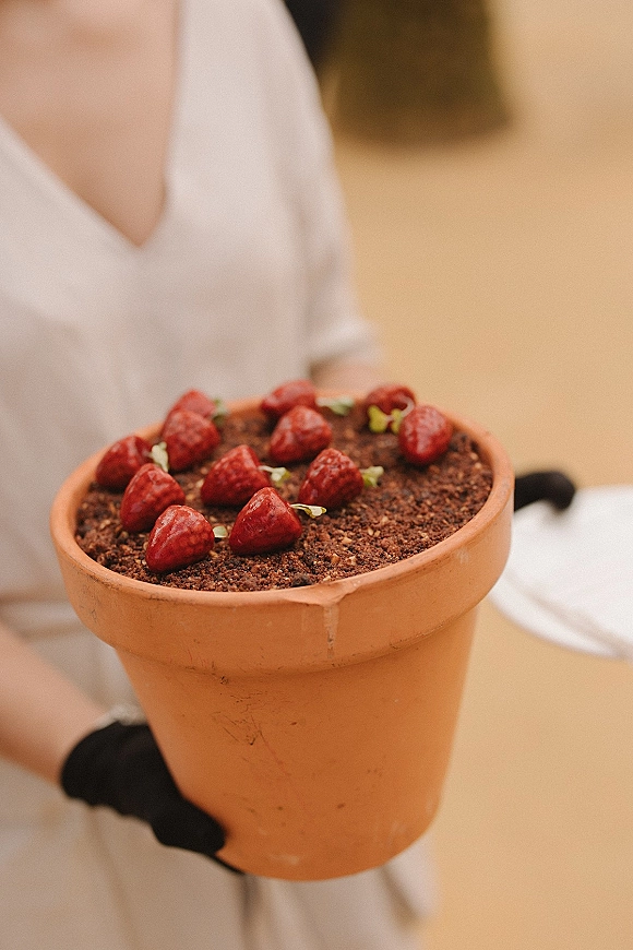 Wedding dessert in a terracotta pot with chocolate crumb topping, strawberries, and microgreens garnish held by black-gloved hands near a white dress