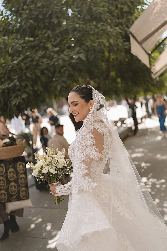 Bridal portrait of a bride holding bouquet of white roses and greenery, looking over her shoulder in a lace mantilla veil on a sunlit walkway