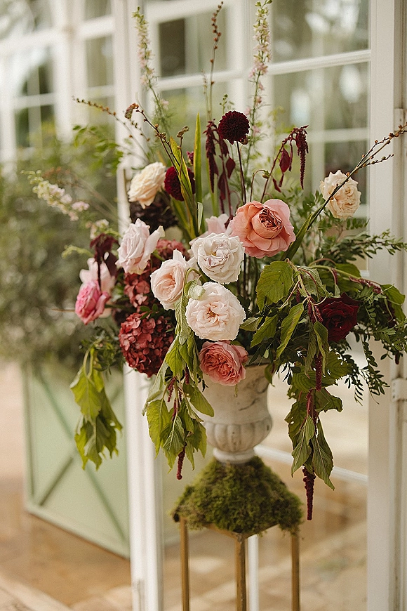 Wedding floral arrangement in a ceramic urn with garden roses, burgundy blooms, and greenery on a pedestal before greenhouse windows