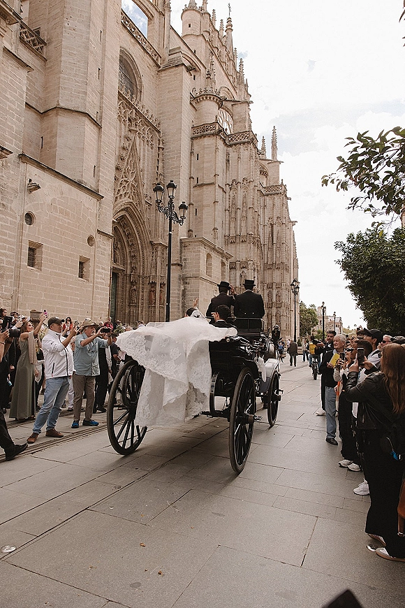 Wedding carriage exit as bride in a horse drawn wedding carriage, veil blowing over her dress train outside a cathedral with cheering crowd