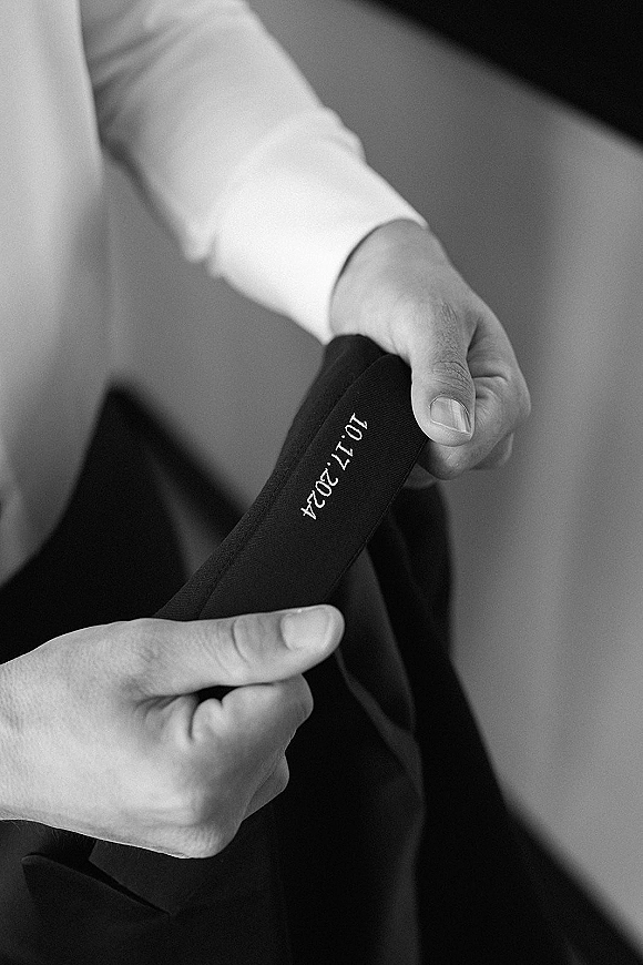 Groom getting ready, putting on a suit jacket with an embroidered wedding date on the lining, hands adjusting over a dress shirt indoors