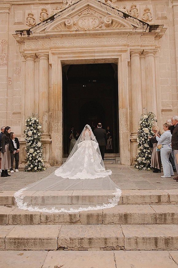 Bride entrance with a cathedral veil and lace wedding dress, walking up stone church steps past white rose florals and guests
